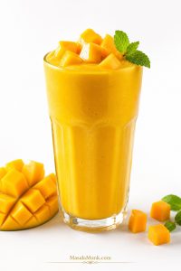 Thick mango smoothie in a glass topped with mango cubes and mint, served with fresh mango and lime.