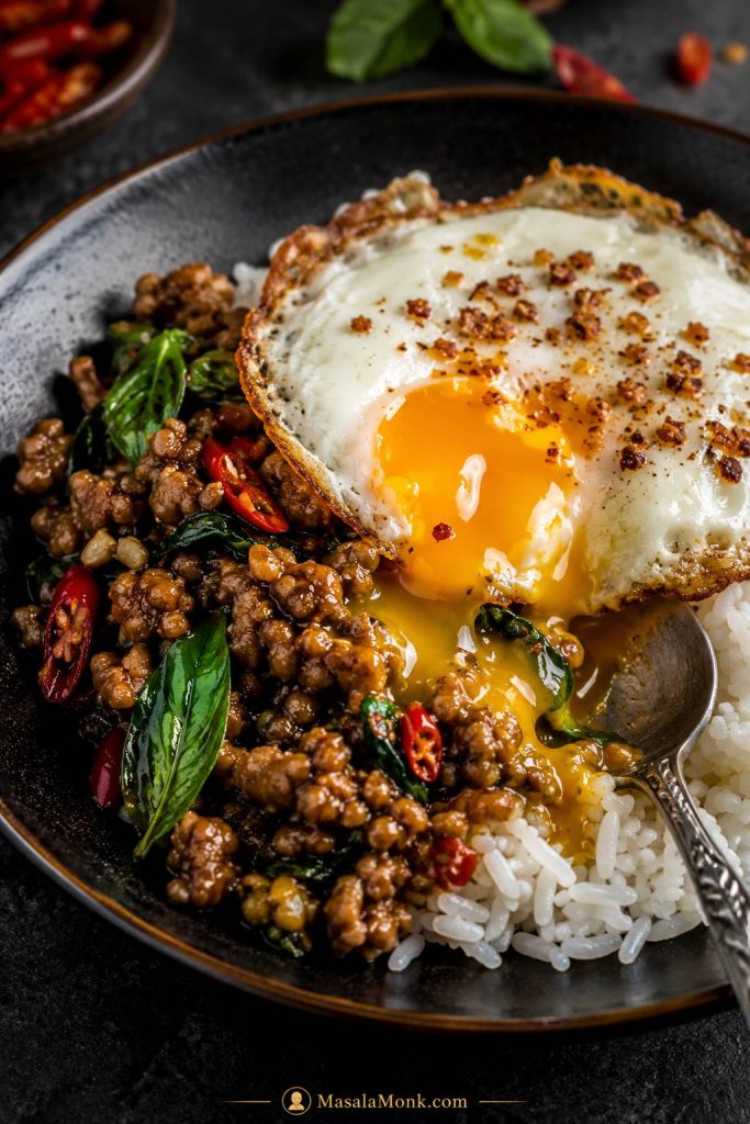 Close-up final Pad Kra Pao serving with glossy basil meat, white rice, crispy fried egg, runny yolk, red chilies, basil leaves, and spoon.