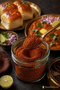 Glass jar of reddish-brown homemade pav bhaji masala with a spoon of spice powder, served beside pav bhaji, buttered pav, onion, lemon, and coriander.