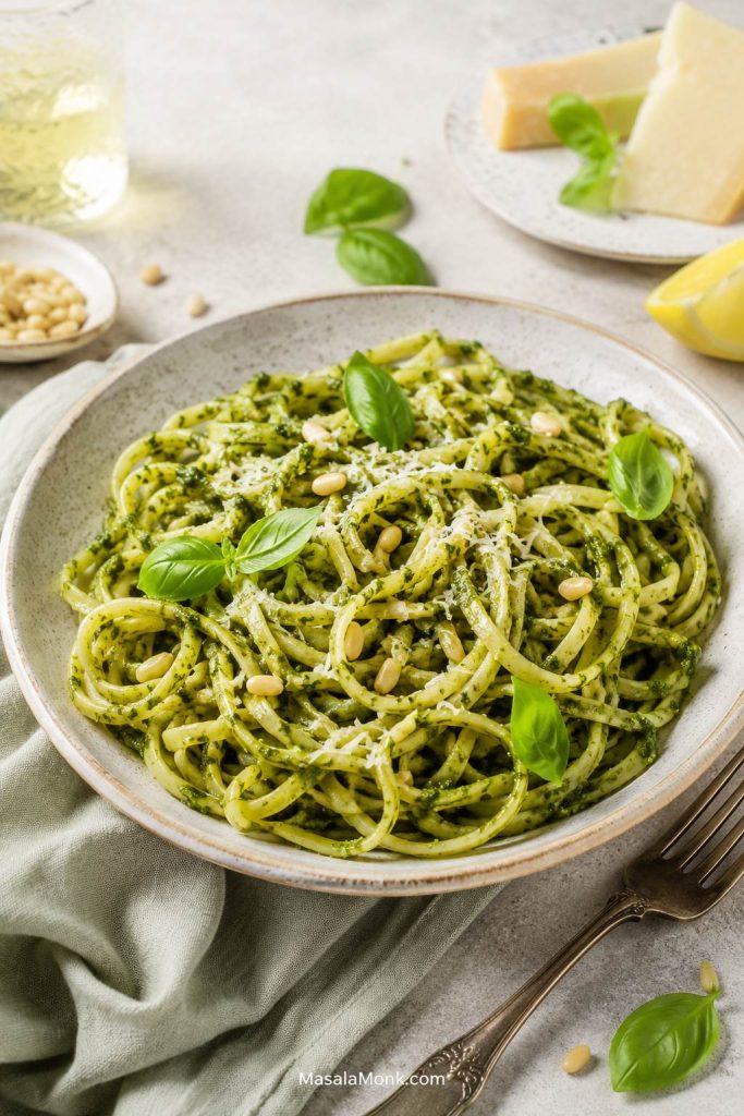Finished pesto pasta in a bowl with basil, Parmesan, pine nuts, lemon and serving utensils