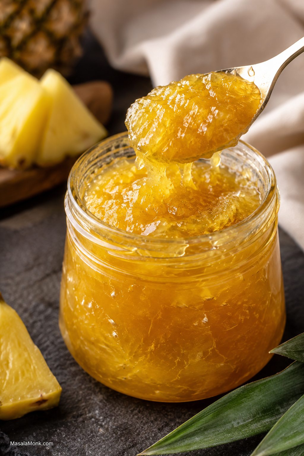 Open glass jar of glossy golden pineapple jam with a spoon lifting thick jam and fresh pineapple wedges behind it