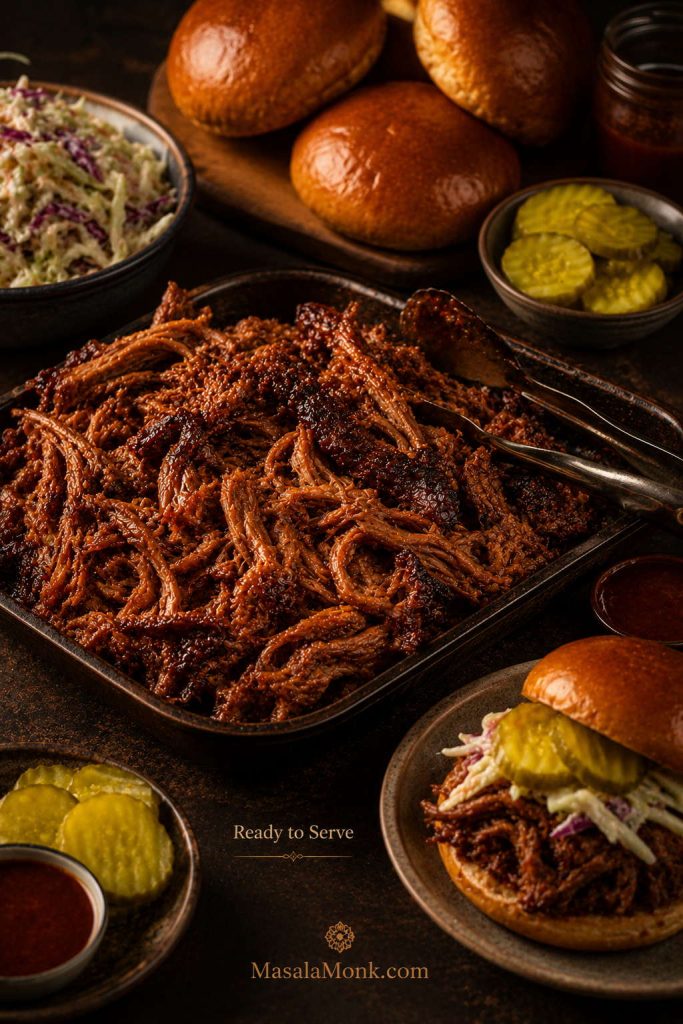 Final serving spread with a tray of glossy pulled pork, buns, slaw, pickles, sauce, tongs, and an assembled pulled pork sandwich.