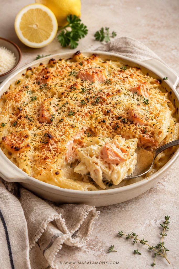 Salmon pasta bake in a cream-colored baking dish with golden Parmesan topping, herbs, and a spoonful lifted out