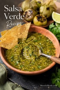 Bowl of roasted salsa verde with tortilla chips, lime, roasted tomatillos, and a spoon showing chunky green texture.