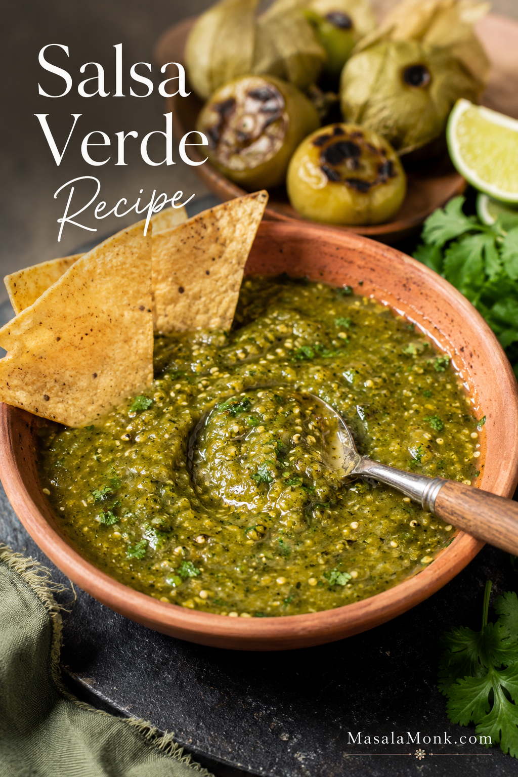 Bowl of roasted salsa verde with tortilla chips, lime, roasted tomatillos, and a spoon showing chunky green texture.