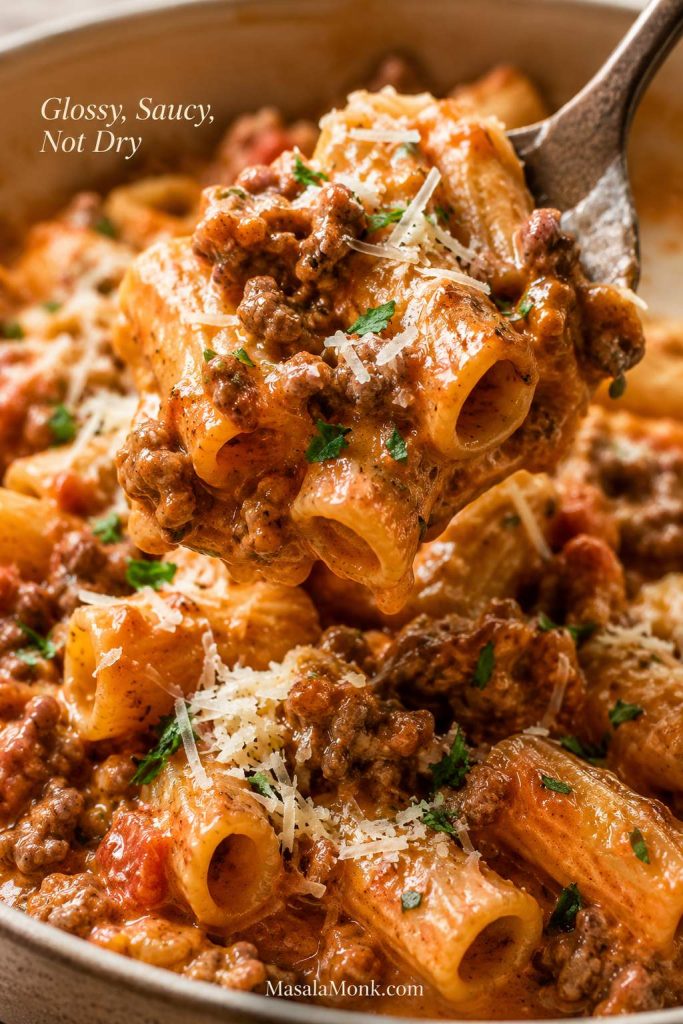 Close-up spoon lift of glossy rigatoni with creamy tomato sauce, crumbled Italian sausage, parmesan, and herbs.