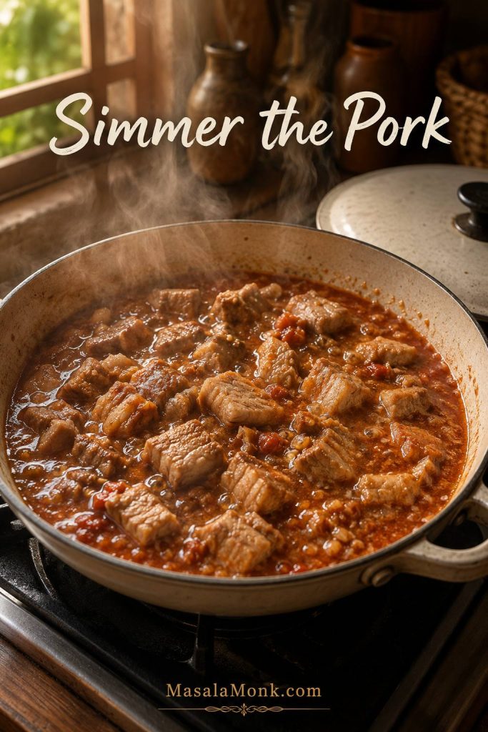 Pork simmering in shallow tomato-bagoong liquid in a wide pan with steam rising.