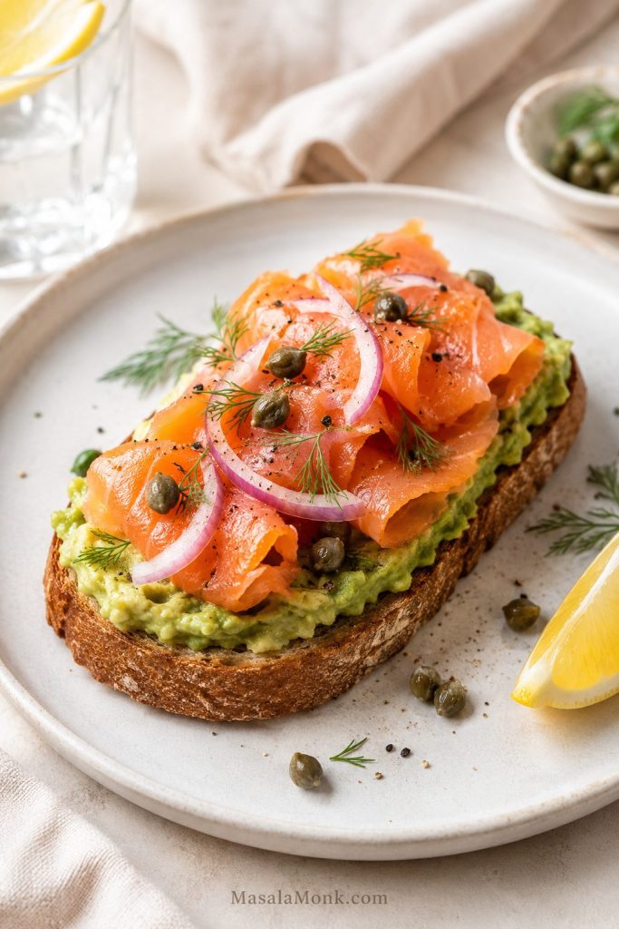 Smoked salmon avocado toast with folded salmon, capers, dill, red onion, lemon, and mashed avocado on sourdough.