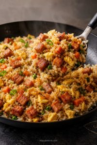 A skillet of fried rice with browned Spam cubes, soft scrambled egg, peas, carrots, scallions, and glossy separated rice being lifted with a spoon.