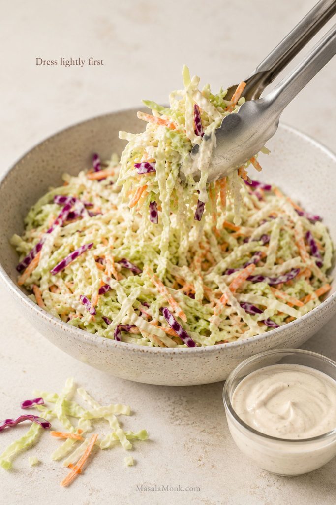 Tongs lifting lightly dressed coleslaw from a bowl with extra dressing set aside.