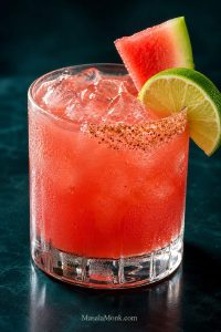 Fresh watermelon margarita on the rocks in a short glass with clear ice, a half-rim, lime wedge, and watermelon garnish.