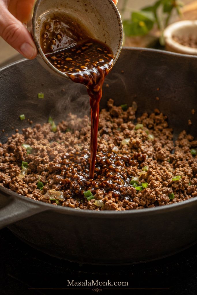 Hand pouring dark soy-garlic sauce from a small bowl into browned ground beef in a skillet.