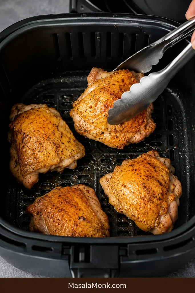 Chicken thighs flipped skin-side up in an air fryer basket with metal tongs.