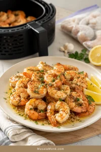Cooked shrimp on a white plate with lemon wedges, garlic butter, parsley, and a blurred air fryer in the background.