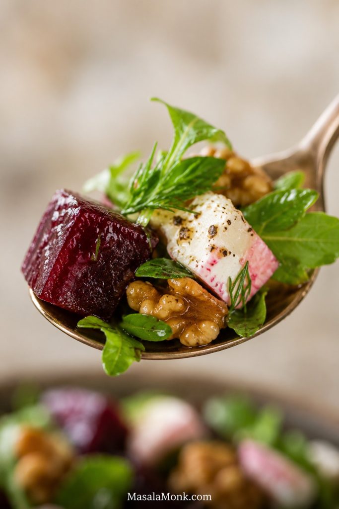 Close-up of a spoon holding roasted beet, feta, walnut, greens, herbs, and glossy dressing.