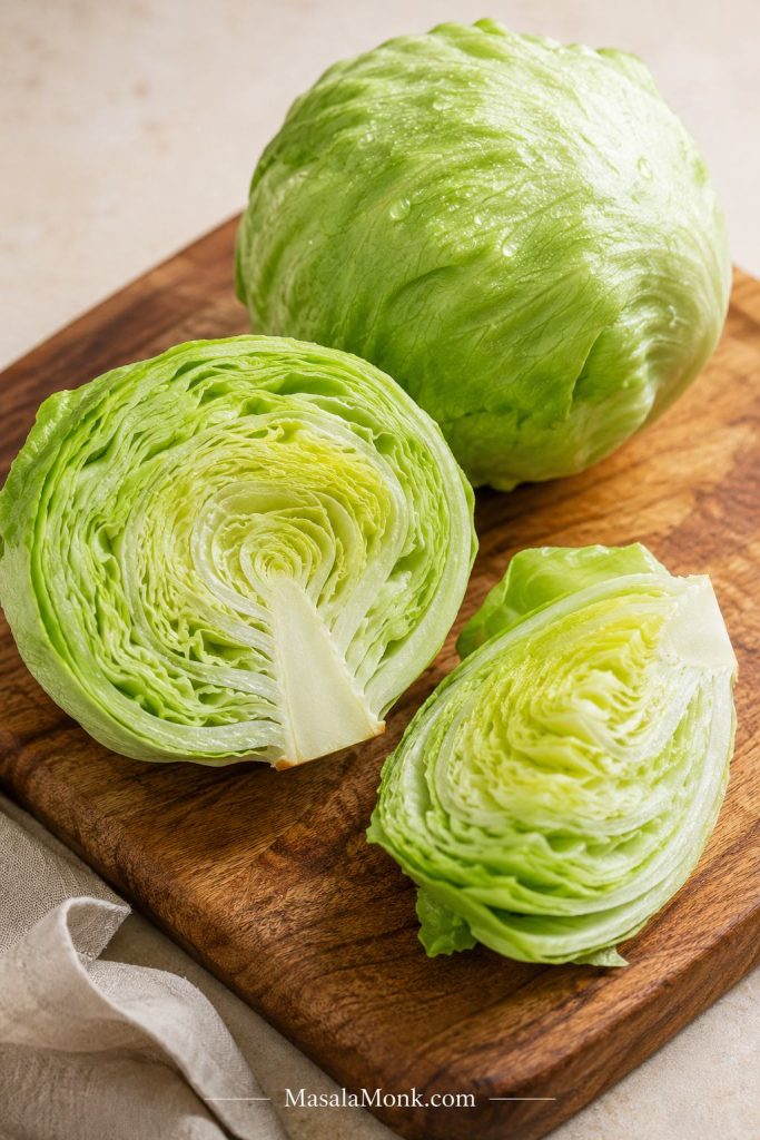 Whole iceberg lettuce, a halved iceberg head, and a quarter wedge on a wooden board showing the tight inner layers and core.