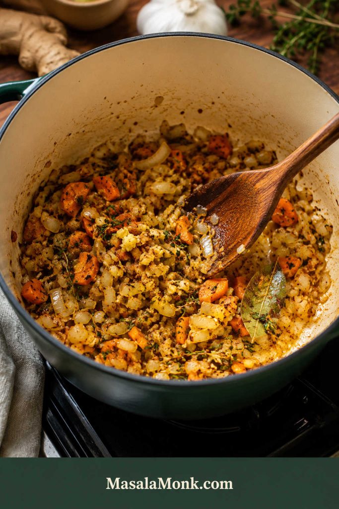 Onion, carrot, garlic, ginger, herbs, spices, and bay leaf sautéing in a green Dutch oven with a wooden spoon.