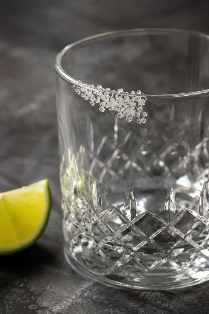 Close-up of a rocks glass with coarse salt on only half the rim and a lime wedge beside the glass.