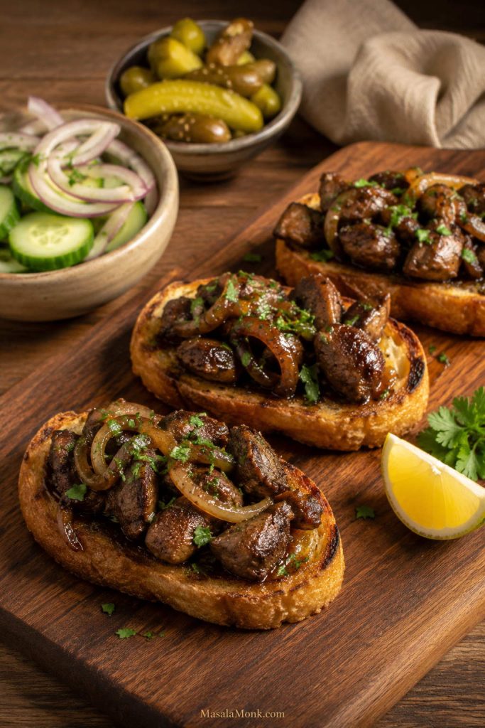 Toasted bread topped with cooked chicken liver pieces, caramelized onions, herbs, and lemon on a wooden board.