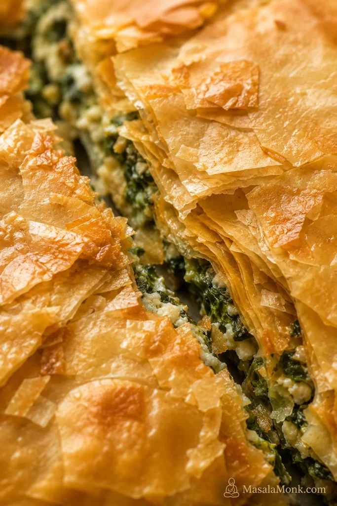A close-up of crisp baked phyllo layers with flaky shards and a glimpse of green filling.