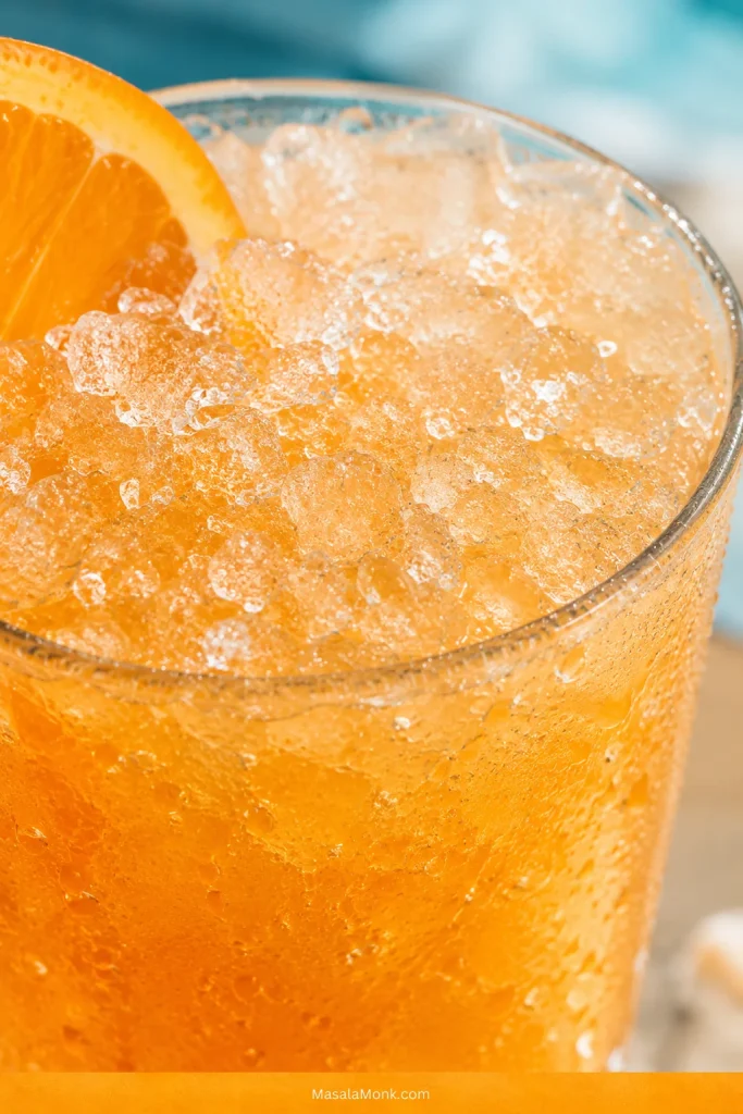 Close-up of crushed ice and condensation in a glass of orange cocktail with an orange slice garnish near the rim.