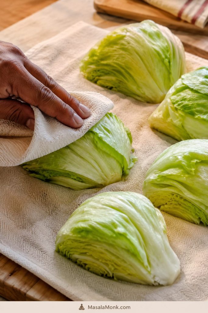 Cut iceberg lettuce wedges drying on a cream towel while a hand pats one wedge dry.