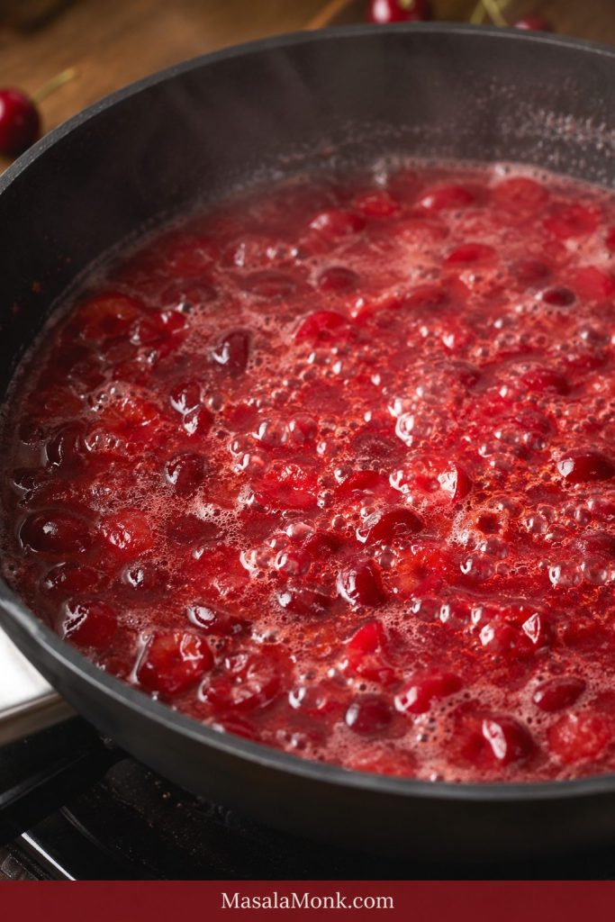 Cherry mixture boiling in a dark pan with thin red liquid, small bubbles, steam, and visible fruit pieces.