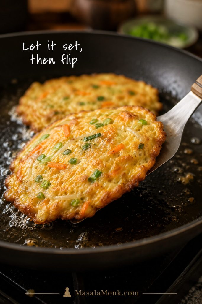Egg Foo Young patty being lifted and flipped on a thin spatula in a skillet with shallow oil.