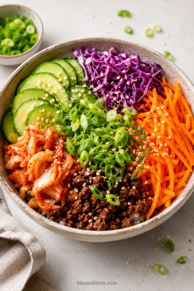 Bright rice bowl with ground beef, sliced cucumber, shredded carrot, purple cabbage, kimchi, scallions, and sesame seeds.