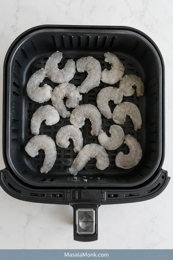 Grey frozen raw shrimp with visible frost spread in a mostly single layer inside a black air fryer basket.