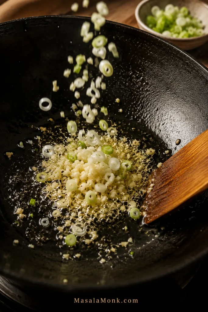 Minced garlic and sliced scallions sizzling in oil inside a black wok with a wooden spatula.