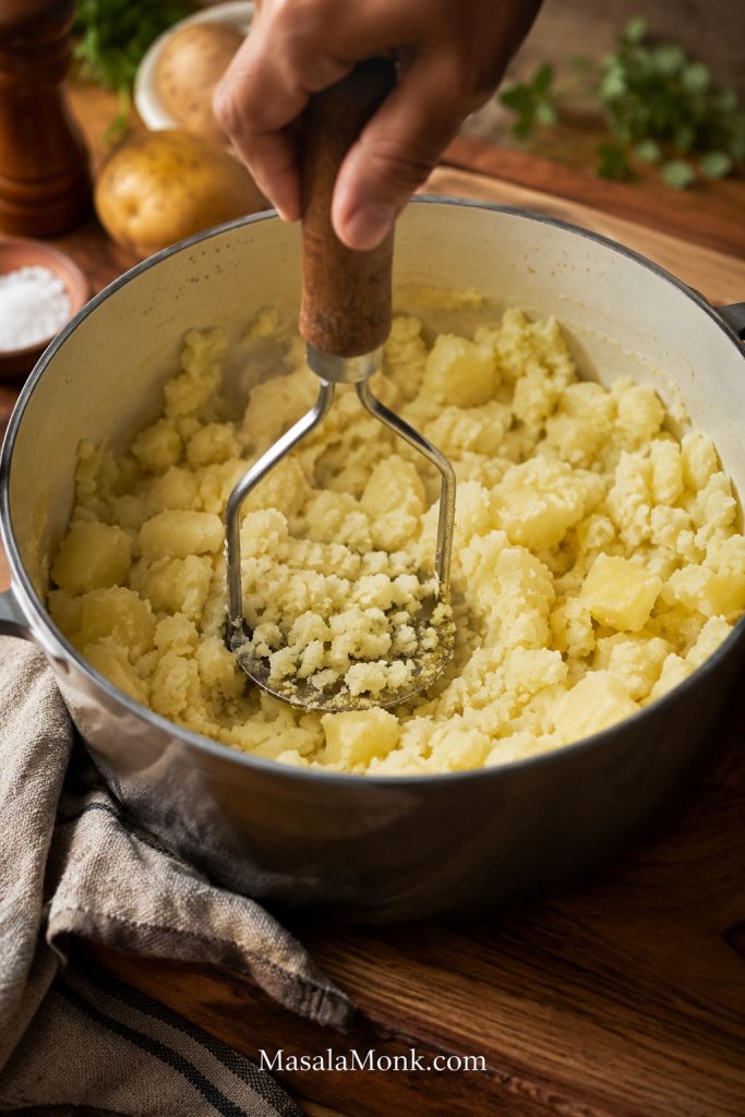 Hand using a potato masher to mash cooked potatoes in a pot.