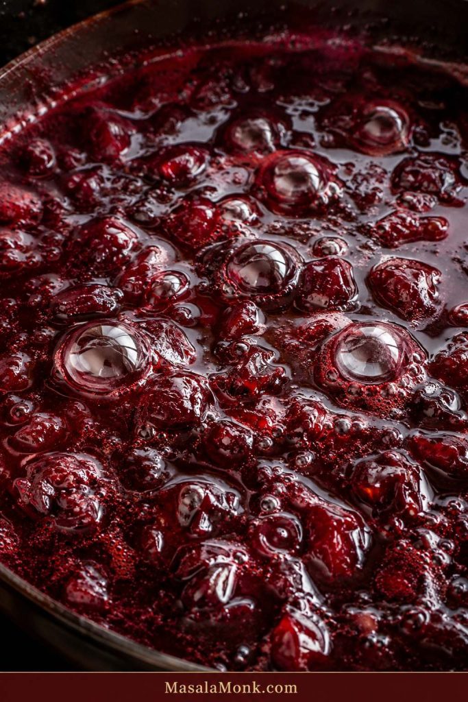 Close-up of thick dark ruby cherry jam bubbling in a pan with shiny syrup and pieces of cherry.