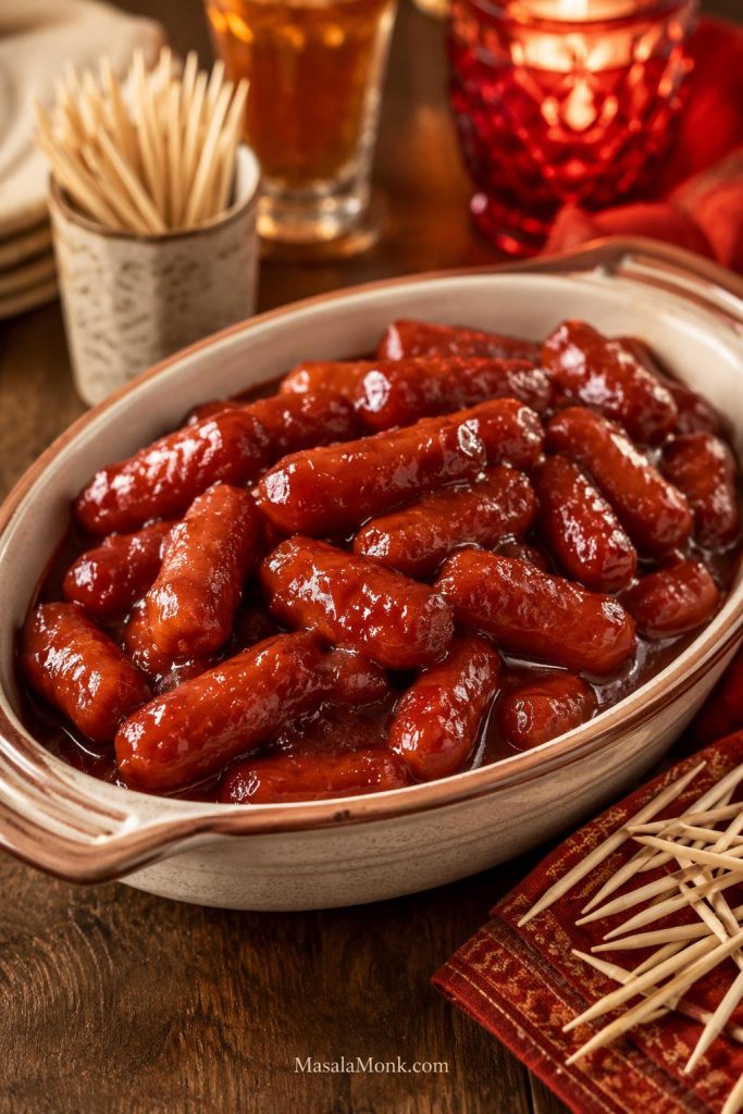 Little Smokies cocktail sausages coated in glossy grape jelly chili sauce in a shallow bowl with toothpicks nearby.