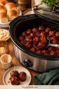 Slow cooker filled with glossy red-brown meatballs, with a serving spoon, toothpicks, small plates, slider buns, and folded napkins on a party table.
