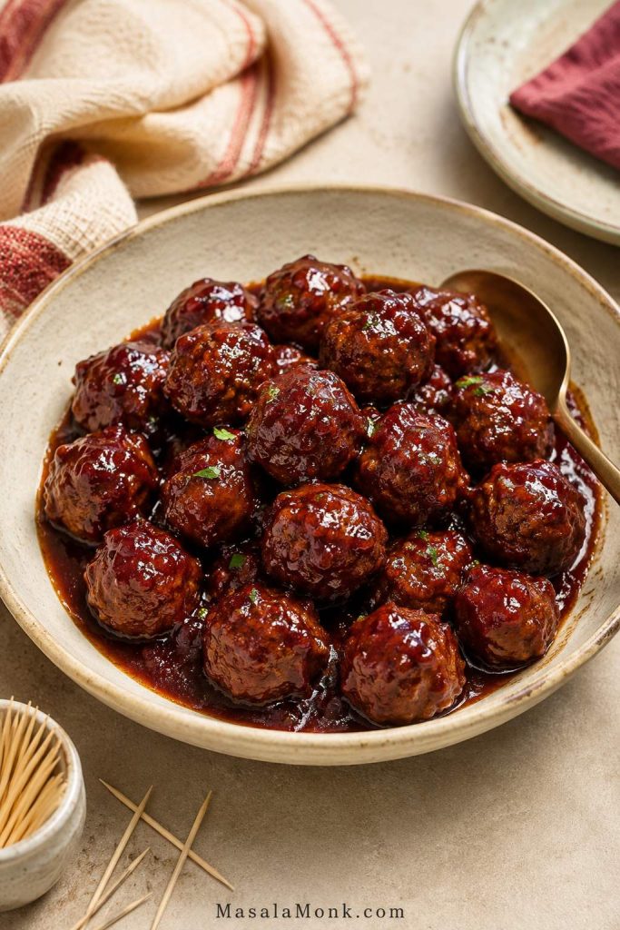 Finished grape jelly meatballs in a serving bowl with sauce, toothpicks, a spoon, folded napkins, and a small plate.