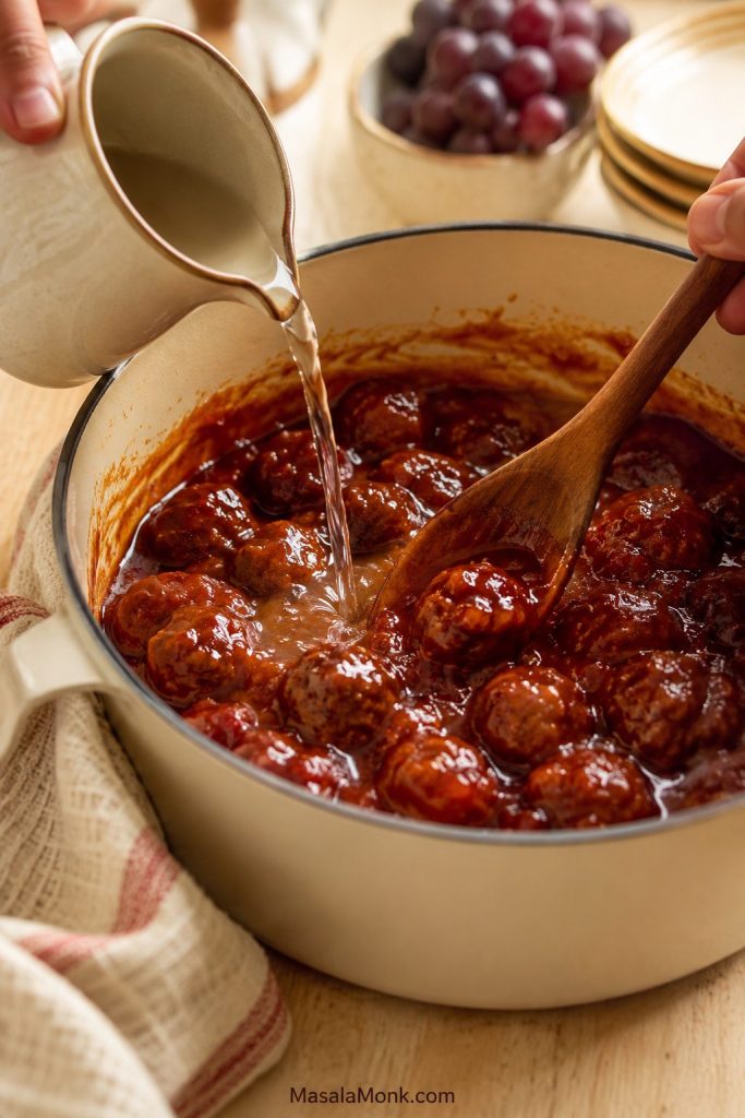 Clear liquid being poured into thick grape jelly meatball sauce while a wooden spoon stirs the meatballs.