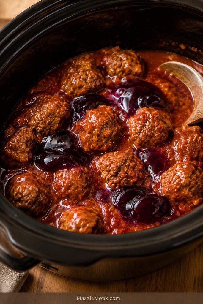 Grape jelly melting into chili sauce around cooked meatballs in a black slow cooker.