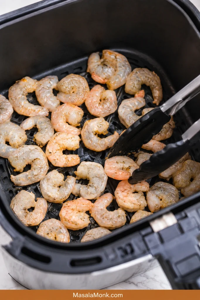 Partly cooked shrimp in an air fryer basket with tongs turning the pieces halfway through cooking.