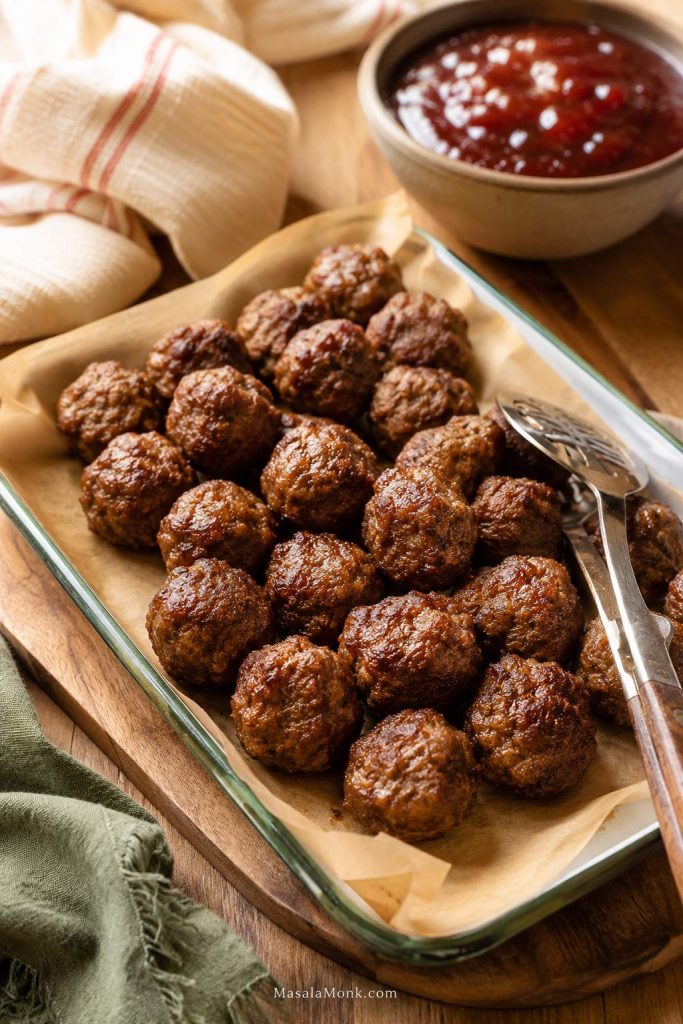 Cooked homemade meatballs on parchment paper in a glass baking dish with tongs and a bowl of red sauce in the background.