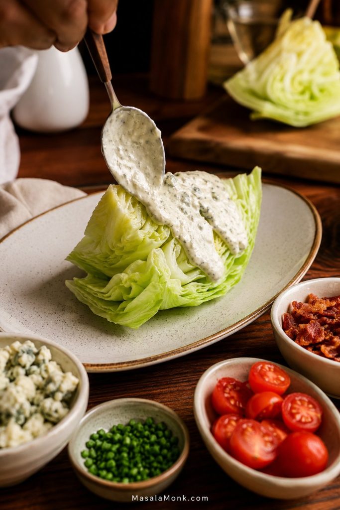 Hand spooning thick blue cheese dressing over an iceberg wedge before adding bacon, tomatoes, chives, and blue cheese crumbles.