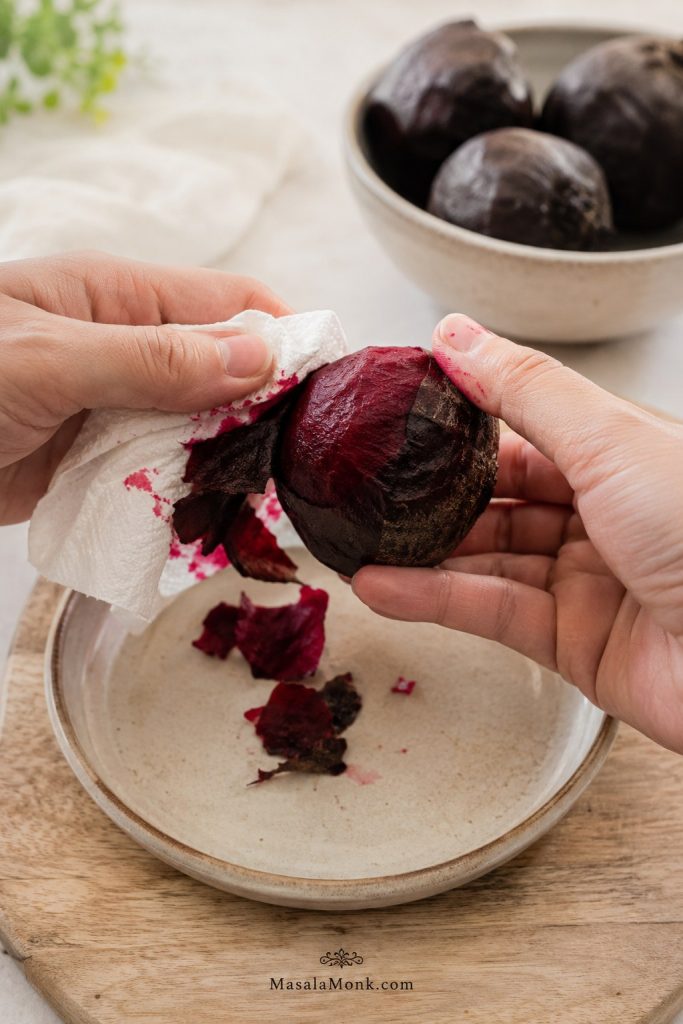 Hands peeling the skin from a roasted beet with a paper towel on a light plate.