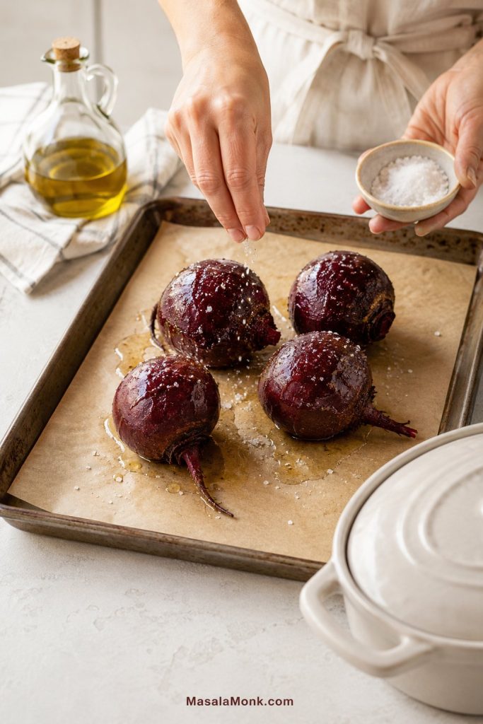 Whole beets on a parchment-lined tray being seasoned with olive oil and salt before roasting.