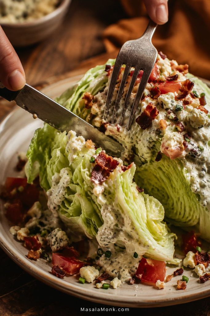 Fork and knife cutting into a dressed iceberg wedge salad with visible lettuce layers, blue cheese dressing, bacon, tomatoes, and crumbles.