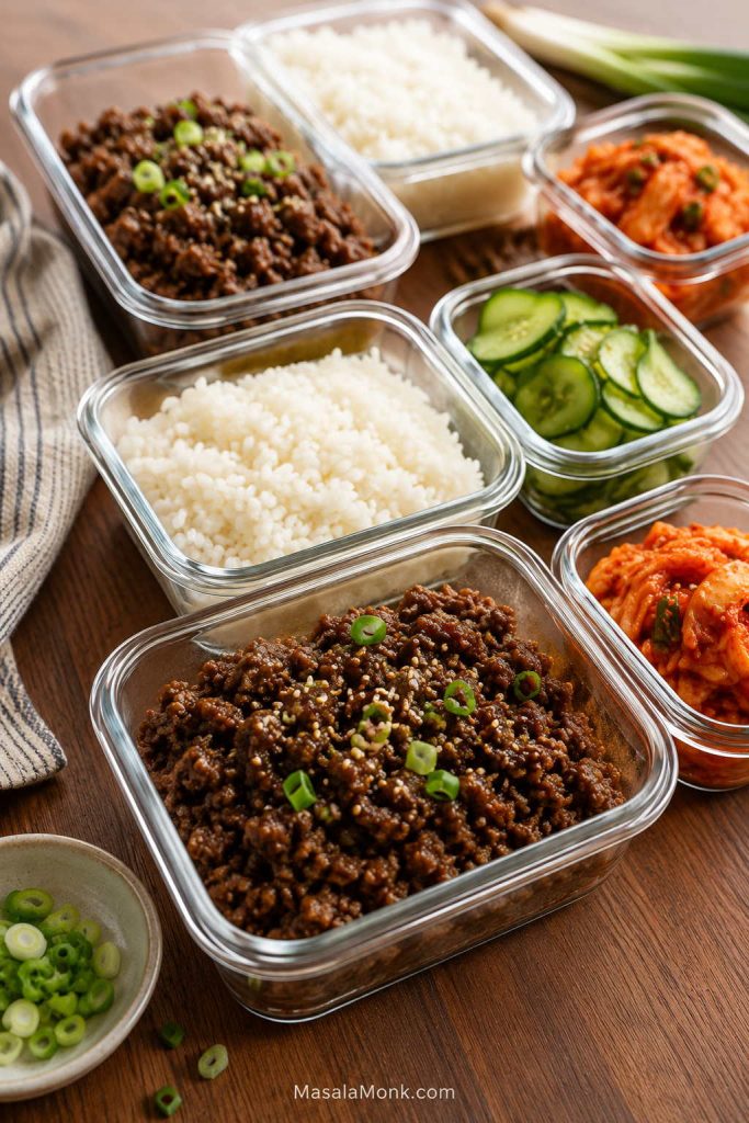 Glass meal prep containers with ground beef, white rice, sliced cucumber, kimchi, scallions, and sesame on a wooden table.