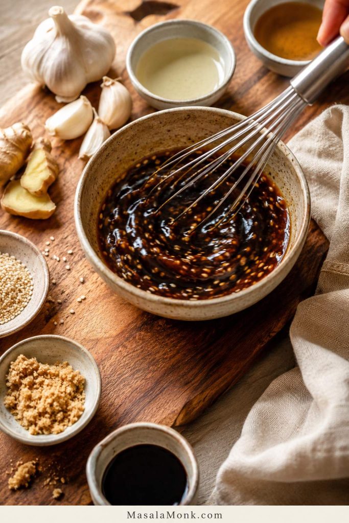 Small ceramic bowl of dark soy-garlic-sesame sauce being whisked, with garlic, ginger, brown sugar, sesame seeds, and sauce ingredients nearby.