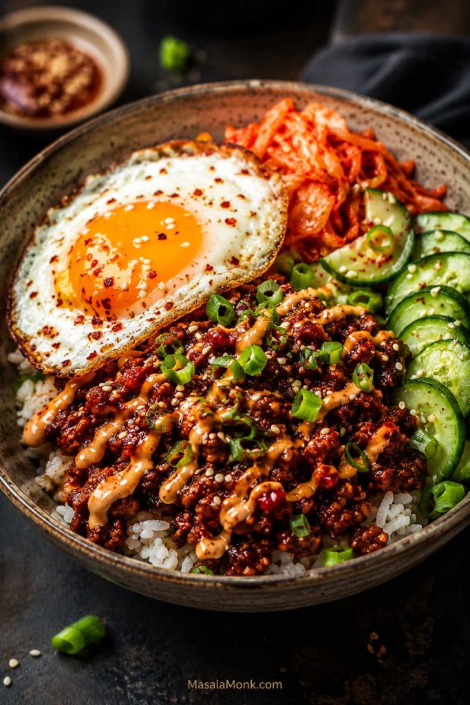 Loaded gochujang beef rice bowl with fried egg, kimchi, cucumber slices, scallions, sesame seeds, creamy drizzle, and white rice.