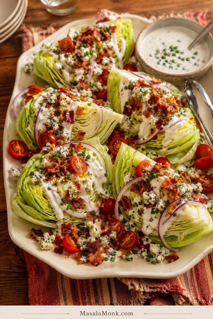 Loaded wedge salad platter with several iceberg wedges, blue cheese dressing, bacon, tomatoes, red onion, chives, blue cheese crumbles, and a dressing bowl.