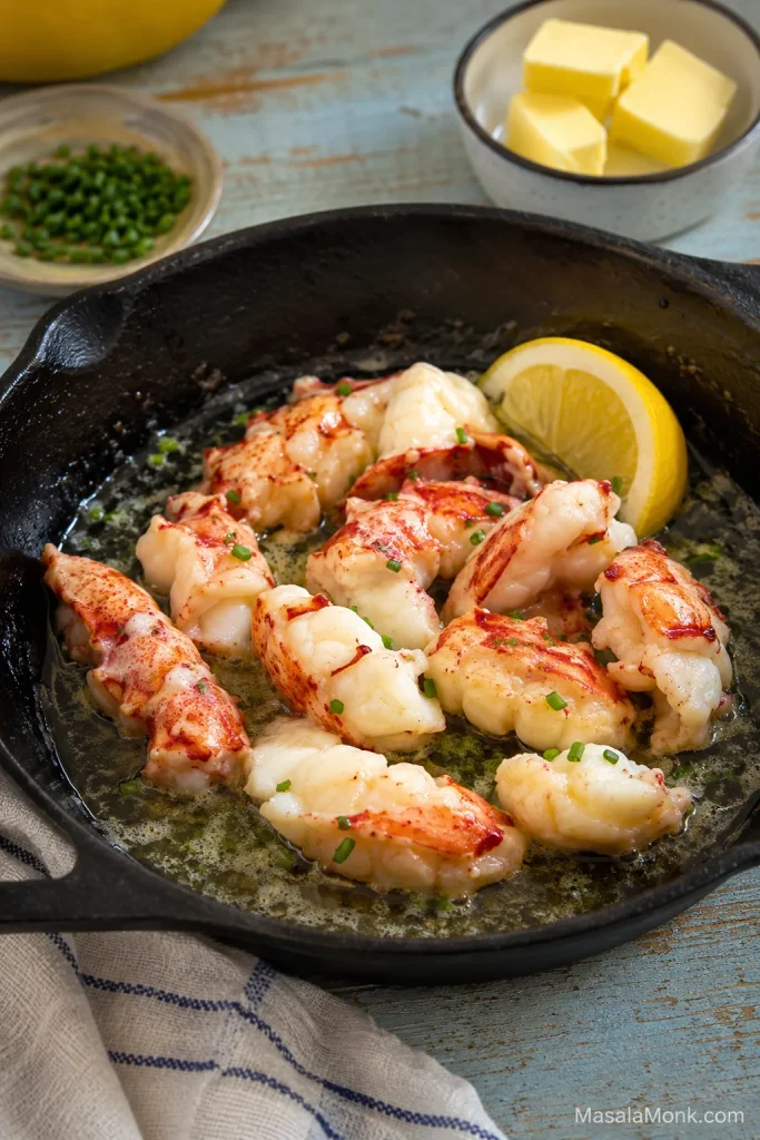 Chunks of cooked lobster meat warming gently in melted butter in a skillet with lemon, chives, and butter nearby.