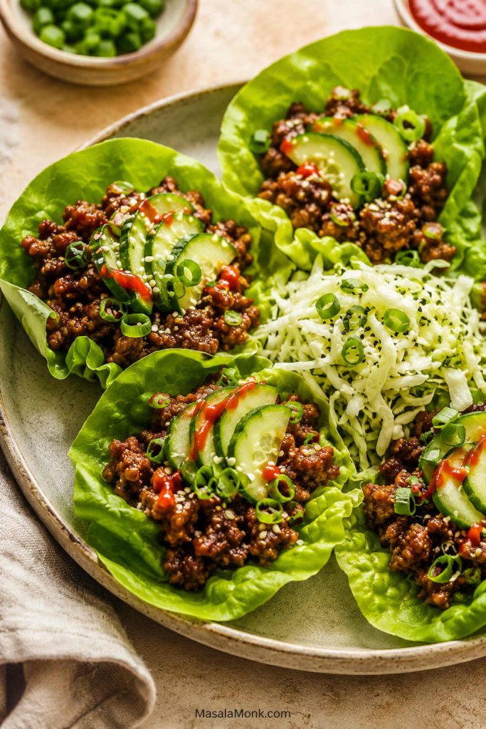 Lettuce cups filled with saucy ground beef, cucumber slices, scallions, sesame seeds, shredded cabbage, and a small red sauce drizzle.